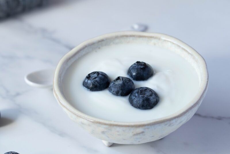 on a marbled surface is a white rustic bowl with yogurt with fresh blueberries on top, behind it is a ceramic spoon