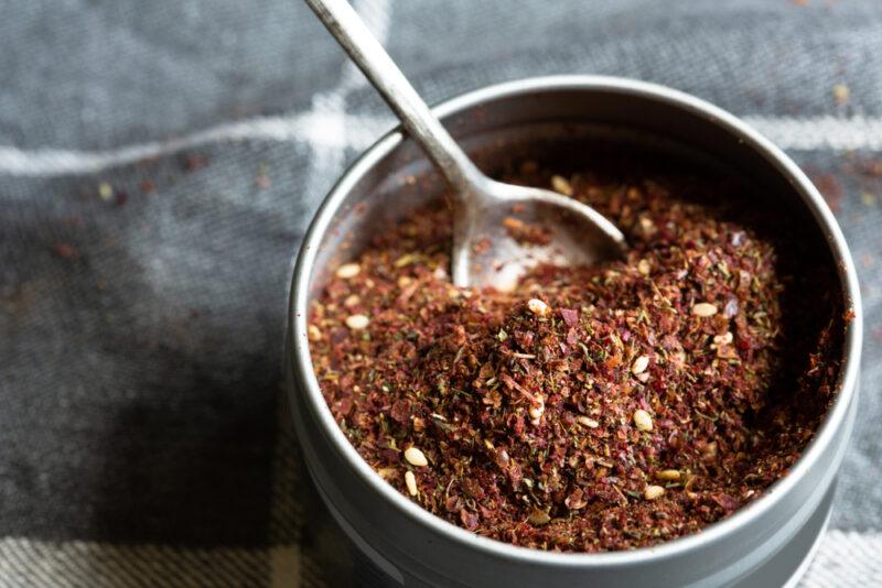 on a gray-colored table napkin with white lines is a top view image of a metal round container with za'atar and a little metal teaspoon