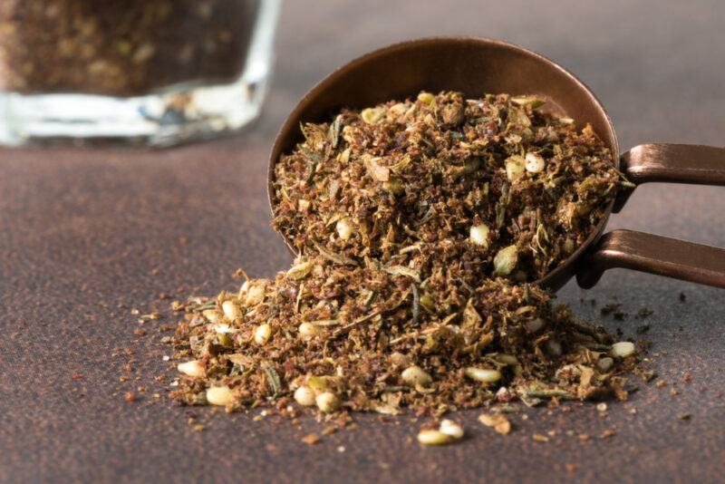 on a dark surface is a metal measuring spoon full of za'atar with some of it spilling over, at the back is a partial image of a clear glass jar with za'atar