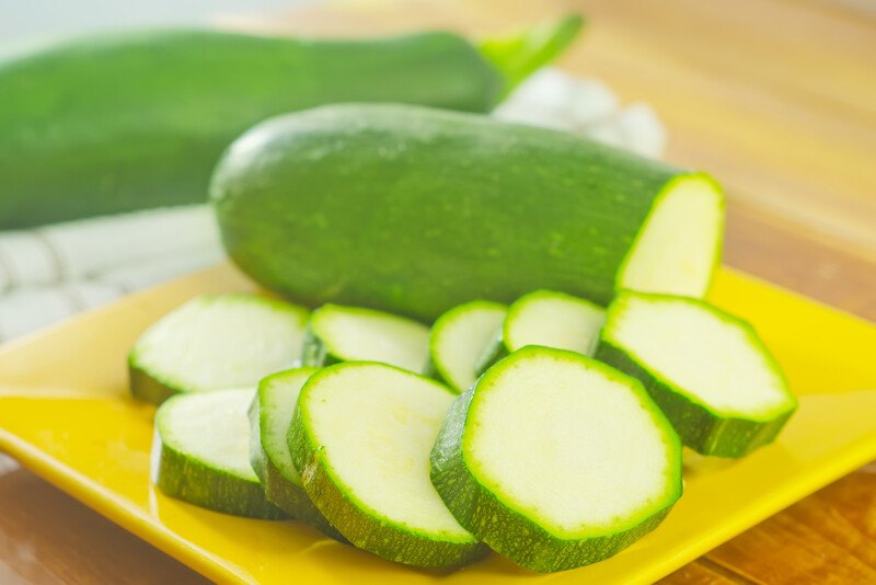 on a wooden surface is a closeup image of zucchini slices on a yellow square dish with whole zucchini at the back