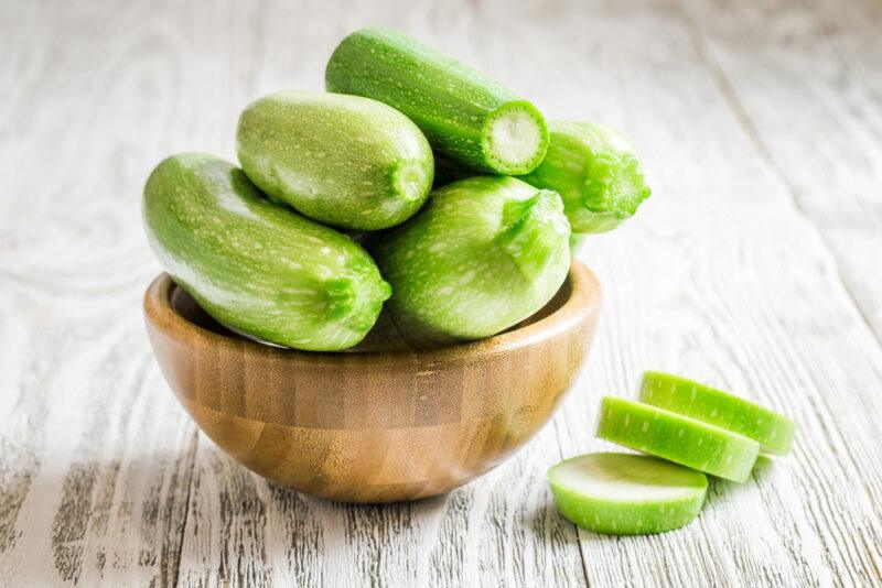 on a wooden surface is a wooden bowl with a pile of whole zucchinis, beside it are three zucchini slices