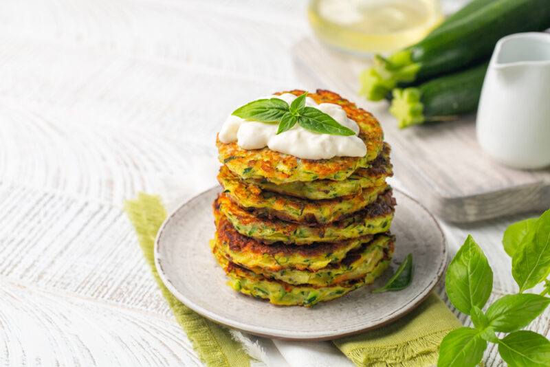 on a white wooden surface is a light grey plate with a pile of zucchini fritters with white sauce and herb on top, resting on a green table napkin with a sprig of herb beside it, at the back are a couple of fresh zucchinis on a wooden chopping board