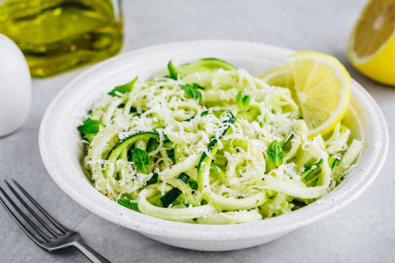 a closeup image of a white shallow bowl of zucchini noodles with parmesan, herb, and a slice of lemon on top, around it is a silver for, a clear jar of olive oil, and a halve lemon